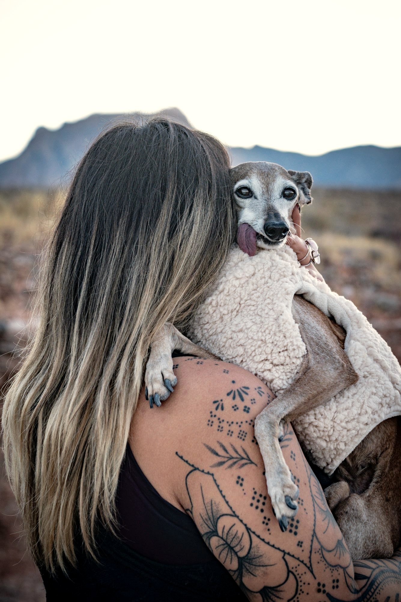 Xsana Saphire holding Bella, her Italian greyhound, wrapped in a sherpa vest in the desert at dusk