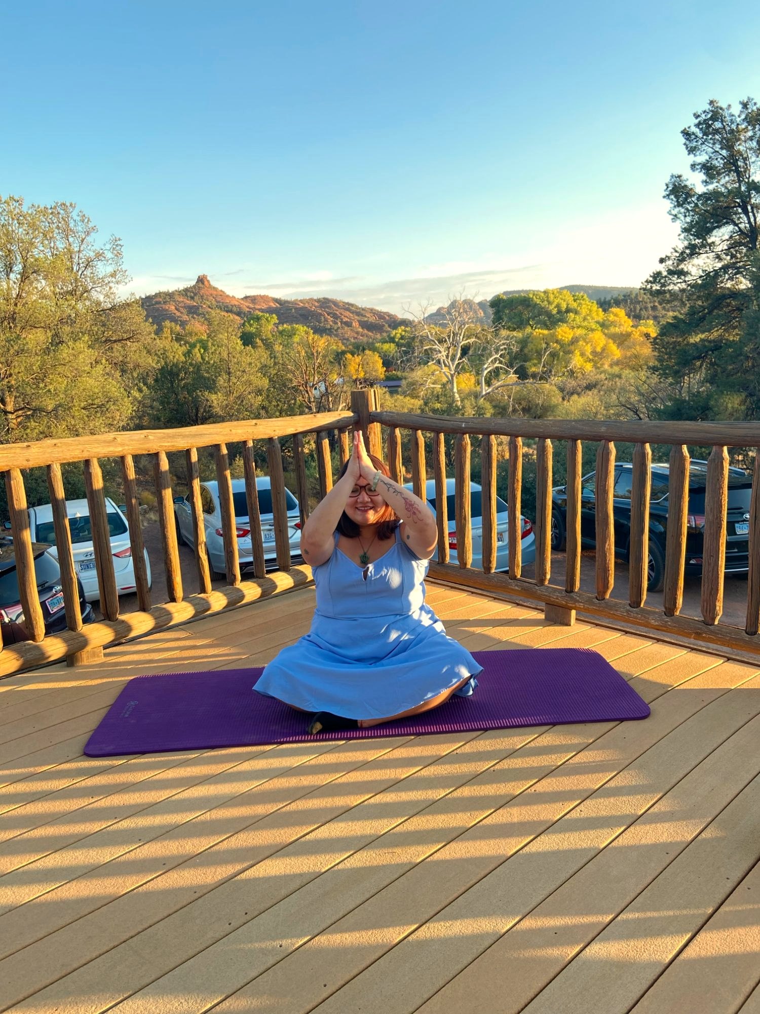 Xsana Saphire seated on a yoga mat on a wooden deck, hands pressed overhead, red rocks behind