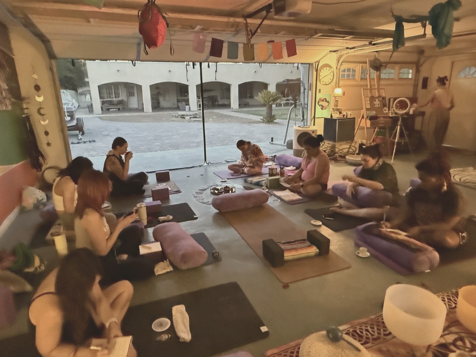 A women's circle at golden hour — heads bowed over journals on mats and bolsters, prayer flags overhead, the evening light coming in.