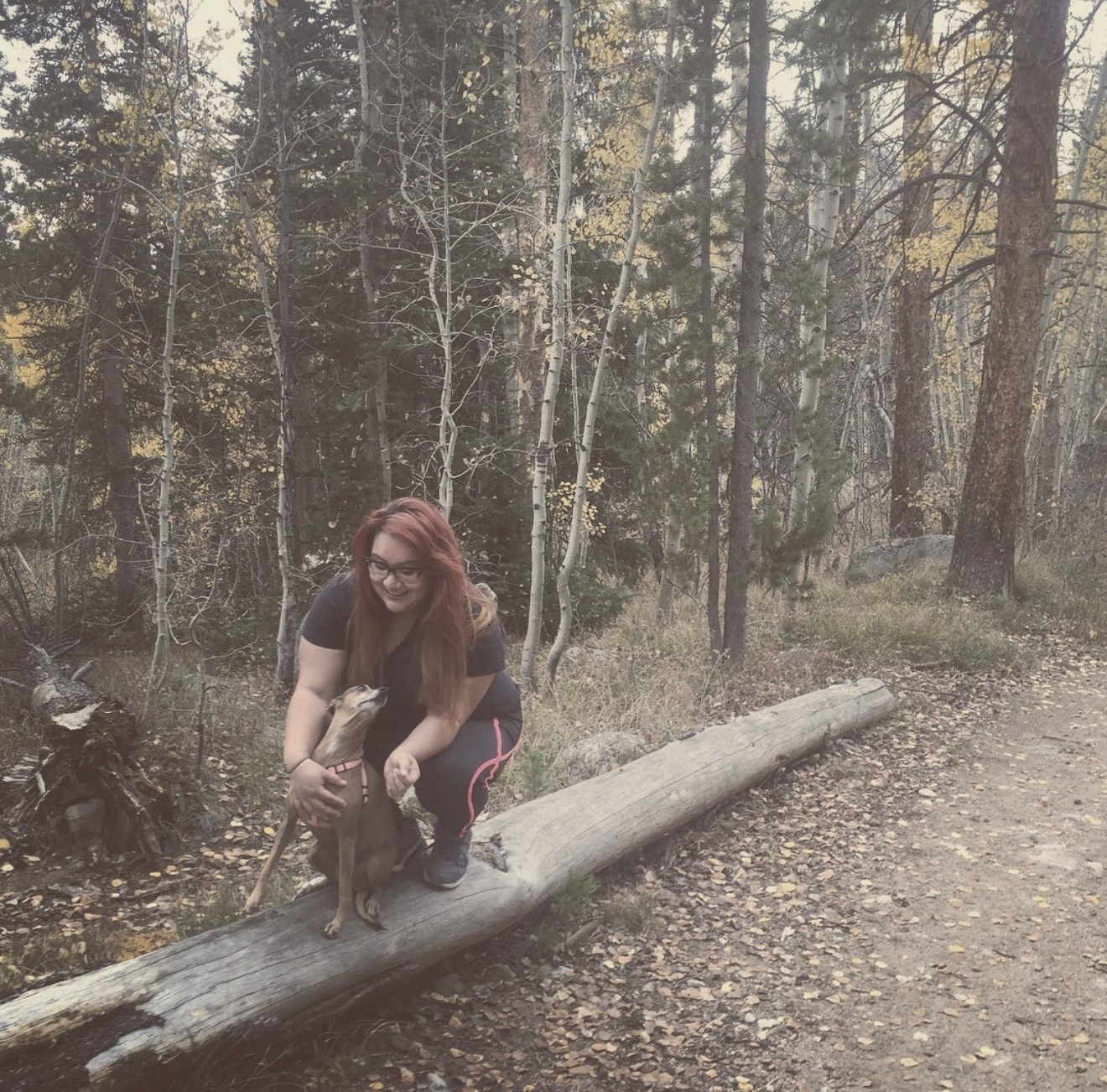 Xsana Saphire with her small dog on a fallen log in a golden Colorado aspen forest.