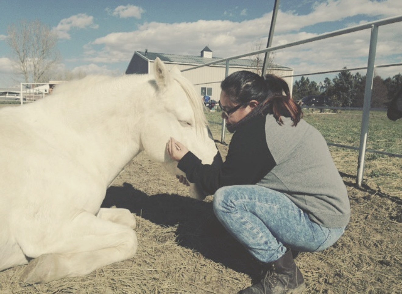 Xsana Saphire kneeling in a corral, forehead resting against a reclining white horse — a wordless, trusting moment of embodied connection.