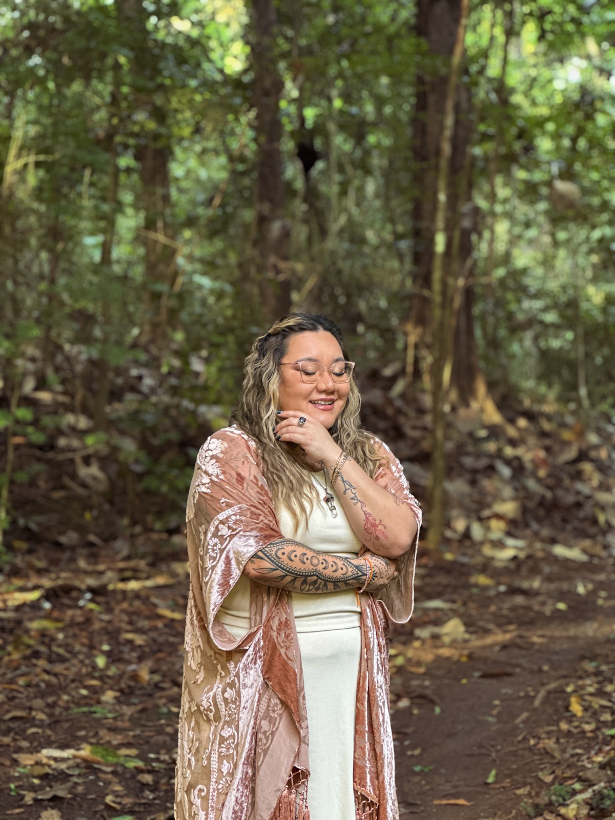 Xsana Saphire standing quietly on a forest path, eyes soft, arms gently crossed — settled and present