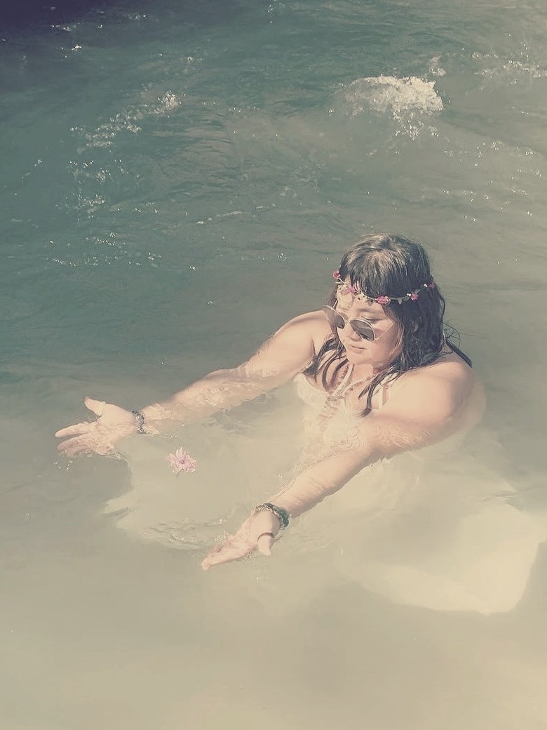 Xsana resting in a milky-turquoise mineral pool wearing a flower crown, releasing a single bloom onto the still water — embodiment as sensation, not story.